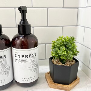 May include: Two brown glass bottles with black pump tops and white labels. The labels read "No. 7 Cypress hand lotion 8 fl oz / 237 ml" and "Magnolia Home". The bottles are on a white tiled surface next to a black hexagonal planter with a small green plant.