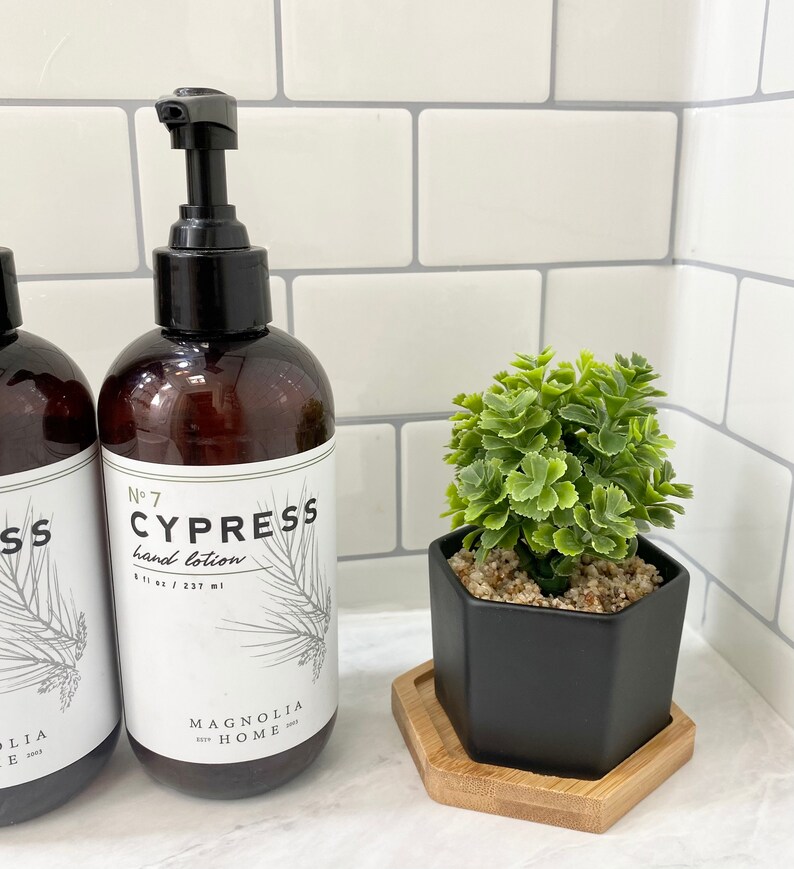 May include: Two brown glass bottles with black pump tops and white labels. The labels read "No. 7 Cypress hand lotion 8 fl oz / 237 ml" and "Magnolia Home". The bottles are on a white tiled surface next to a black hexagonal planter with a small green plant.