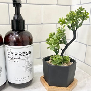 May include: A brown glass bottle with a black pump dispenser. The bottle is labeled "No. 7 CYPRESS hand wash 8.1 fl oz / 237 ml" and "MAGNOLIA HOME". The bottle is next to a black hexagonal planter with a small green plant inside. The planter is on a wooden coaster.