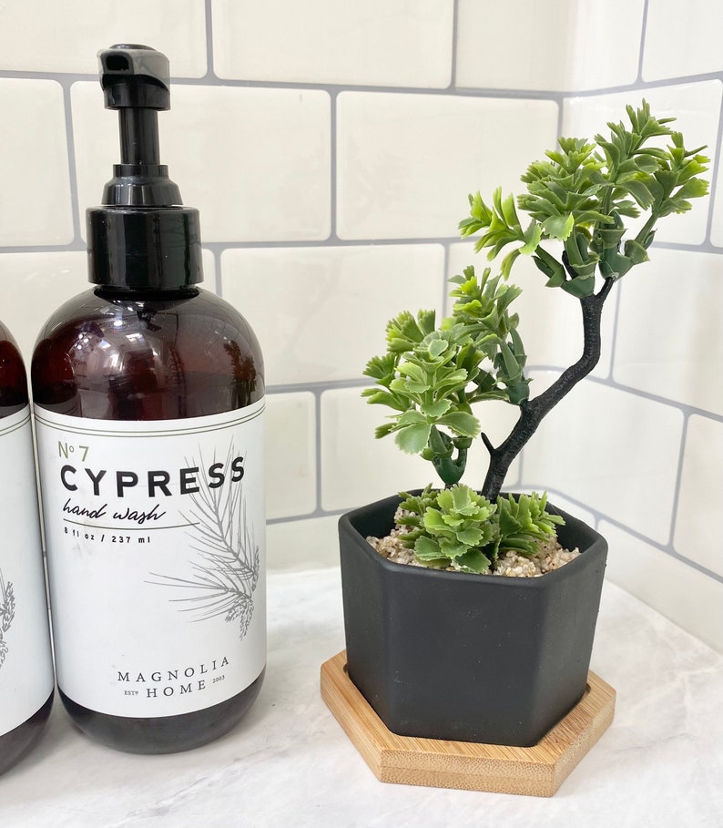 May include: A brown glass bottle with a black pump dispenser. The bottle is labeled "No. 7 CYPRESS hand wash 8.1 fl oz / 237 ml" and "MAGNOLIA HOME". The bottle is next to a black hexagonal planter with a small green plant inside. The planter is on a wooden coaster.