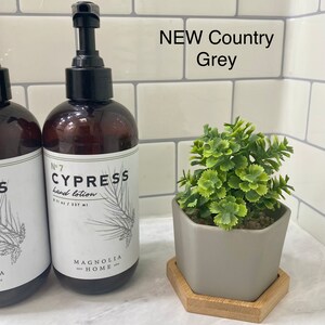 May include: A brown glass bottle of hand lotion with a white label that says "No. 7 Cypress hand lotion 8 fl oz / 237 ml Magnolia Home". The bottle is on a white tiled surface with a small gray hexagonal planter with a green plant on a wooden hexagonal coaster.