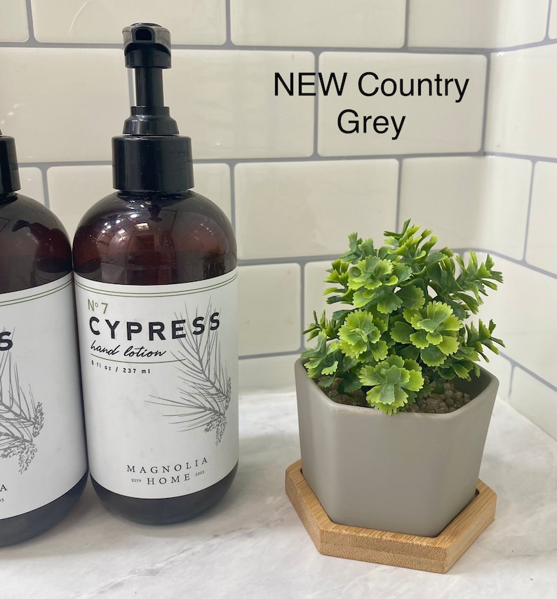 May include: A brown glass bottle of hand lotion with a white label that says "No. 7 Cypress hand lotion 8 fl oz / 237 ml Magnolia Home". The bottle is on a white tiled surface with a small gray hexagonal planter with a green plant on a wooden hexagonal coaster.