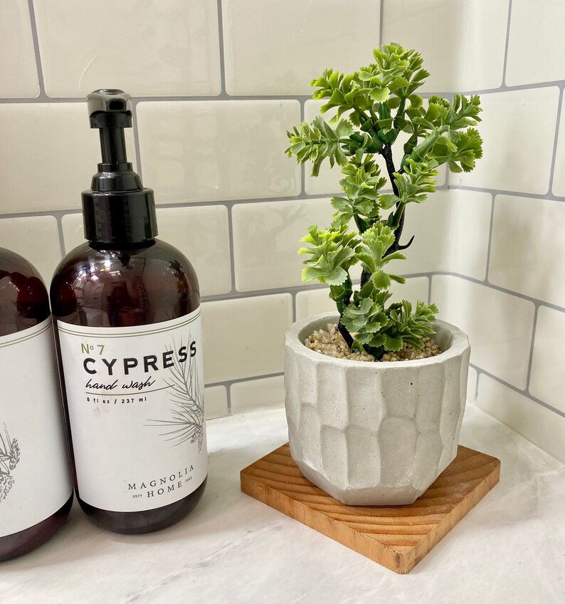 May include: A brown glass bottle with a black pump dispenser labeled "No. 7 CYPRESS hand wash 8 fl oz / 237 ml MAGNOLIA HOME". The bottle is next to a small potted plant with green leaves in a white concrete planter on a wooden coaster.