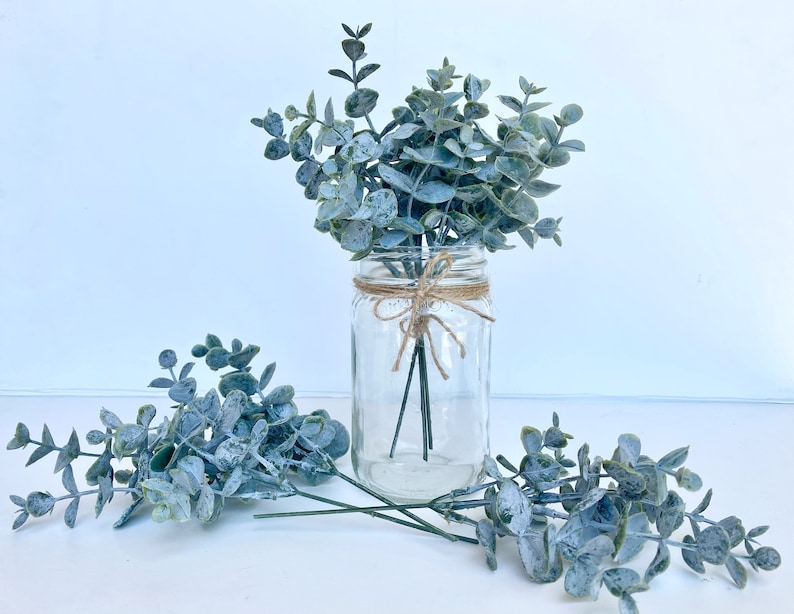 May include: A glass jar with a clear glass lid filled with a bouquet of blue-green eucalyptus stems. The stems are tied together with twine and the jar is sitting on a white surface.