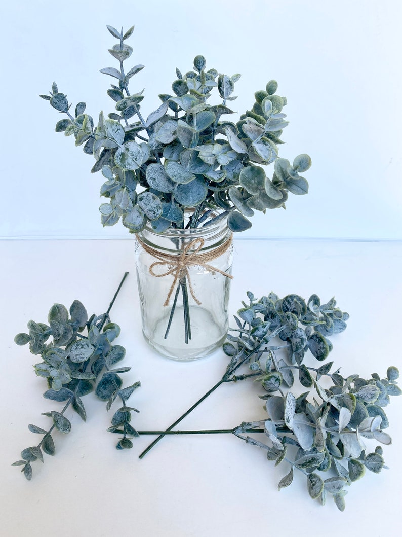 May include: A bouquet of dusty blue eucalyptus stems in a clear glass jar with a brown twine bow. The stems are arranged in a vase on a white surface.