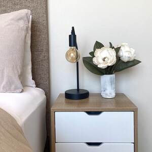 May include: A black metal desk lamp with a round base and a large, exposed lightbulb. The lamp sits on a wooden nightstand with two white drawers. A vase of white flowers with green leaves is also on the nightstand.
