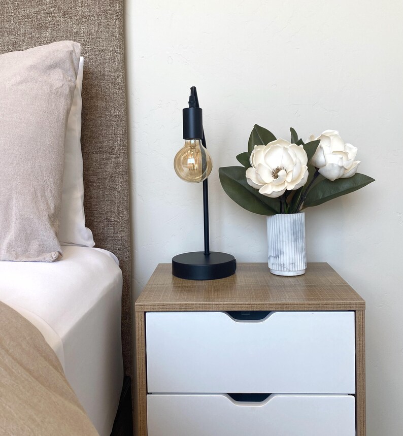 May include: A black metal desk lamp with a round base and a large, exposed lightbulb. The lamp sits on a wooden nightstand with two white drawers. A vase of white flowers with green leaves is also on the nightstand.