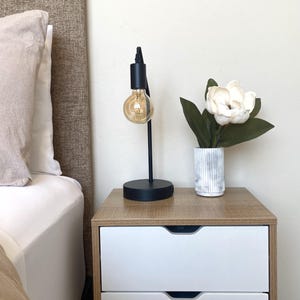 May include: A black desk lamp with a round base and a visible lightbulb sits on a wooden nightstand. A white and gray striped vase holds a white flower with green leaves. A bed with a beige headboard is in the background.