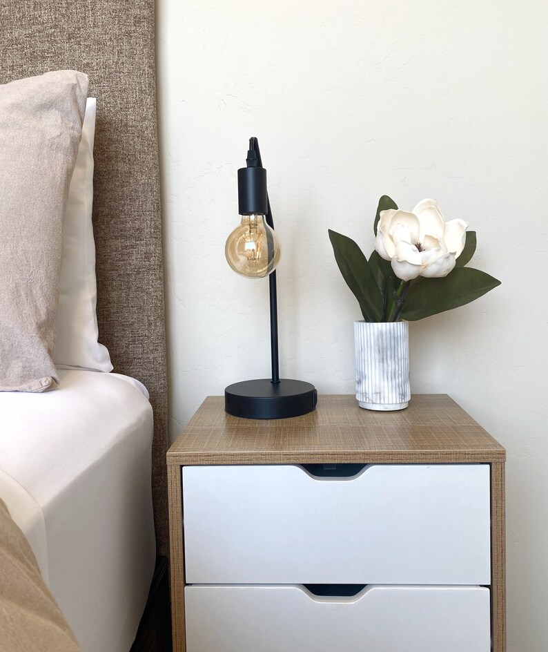 May include: A black desk lamp with a round base and a visible lightbulb sits on a wooden nightstand. A white and gray striped vase holds a white flower with green leaves. A bed with a beige headboard is in the background.