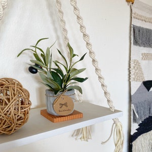 May include: White shelf with a macrame rope hanging system. A small potted plant with a brown tag that says "Handmade" is on the shelf. A woven ball is also on the shelf.