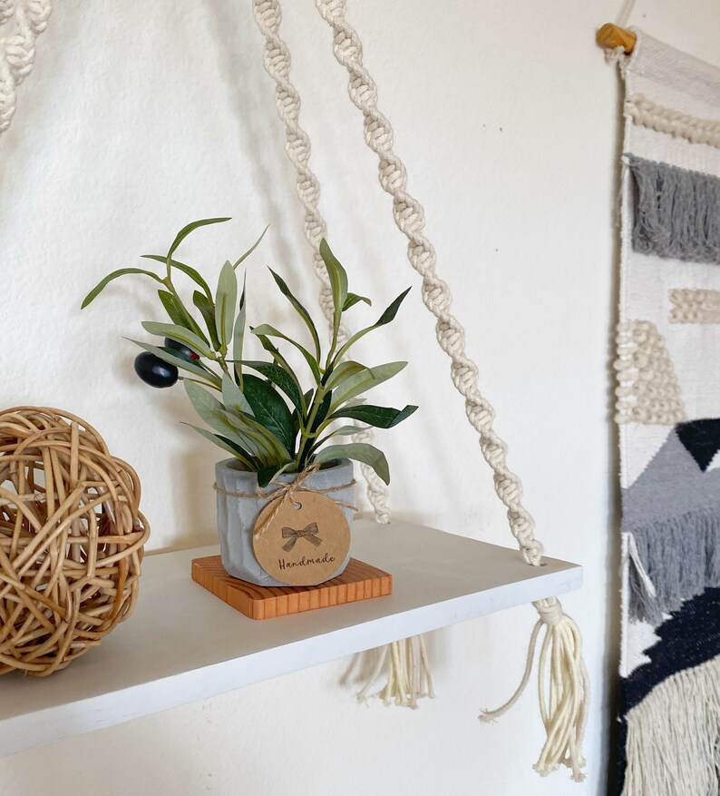 May include: White shelf with a macrame rope hanging system. A small potted plant with a brown tag that says "Handmade" is on the shelf. A woven ball is also on the shelf.