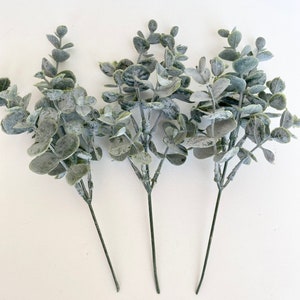 May include: Three sprigs of artificial eucalyptus with a frosted, silvery-green finish. The stems are long and slender, and the leaves are clustered together.