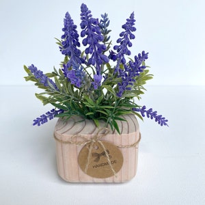 K&ouml;nnte beinhalten: Ein kleiner Holztopf mit einem braunen Etikett, auf dem "HANDMADE" in wei&szlig;en Buchstaben steht. Der Topf ist mit lila Lavendelbl&uuml;ten und gr&uuml;nen Bl&auml;ttern gef&uuml;llt.