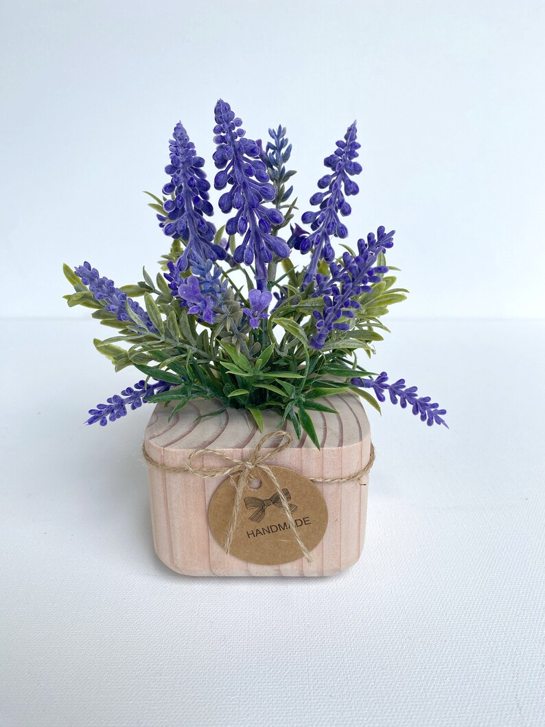 K&ouml;nnte beinhalten: Ein kleiner Holztopf mit einem braunen Etikett, auf dem "HANDMADE" in wei&szlig;en Buchstaben steht. Der Topf ist mit lila Lavendelbl&uuml;ten und gr&uuml;nen Bl&auml;ttern gef&uuml;llt.