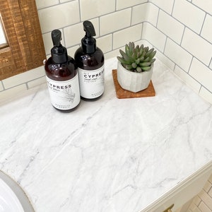 May include: Two brown pump bottles of Cypress hand soap and a small succulent plant in a white pot on a wooden coaster. The bottles have white labels with black text and a botanical illustration. The countertop has a white marble pattern.