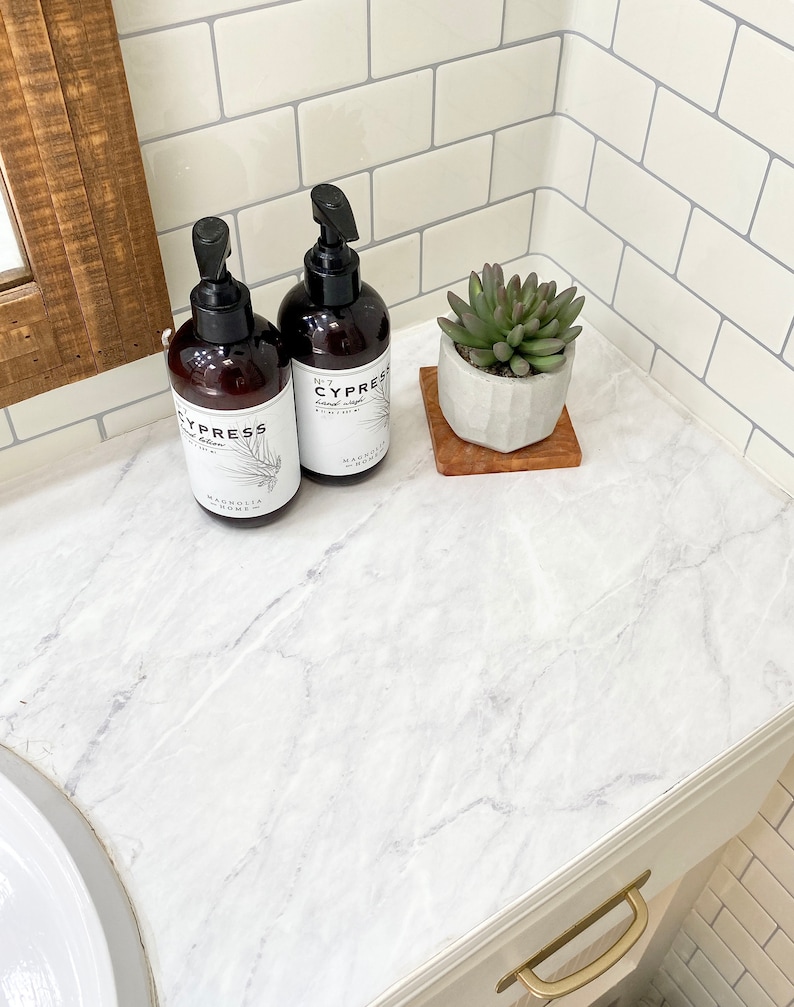 May include: Two brown pump bottles of Cypress hand soap and a small succulent plant in a white pot on a wooden coaster. The bottles have white labels with black text and a botanical illustration. The countertop has a white marble pattern.