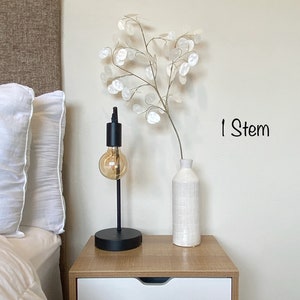 May include: A white ceramic vase with a textured surface holds a single stem of white paper flowers. The vase is on a wooden nightstand with a white drawer. A black lamp with a clear glass bulb is on the nightstand. The text "1 Stem" is written on the wall.