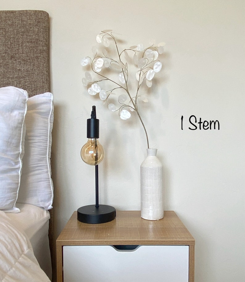 May include: A white ceramic vase with a textured surface holds a single stem of white paper flowers. The vase is on a wooden nightstand with a white drawer. A black lamp with a clear glass bulb is on the nightstand. The text "1 Stem" is written on the wall.