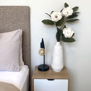 May include: A bedroom nightstand with a black lamp and a white vase holding white magnolia flowers. The nightstand has a light wood top and two white drawers. A bed with a beige headboard and pillow is in the background.