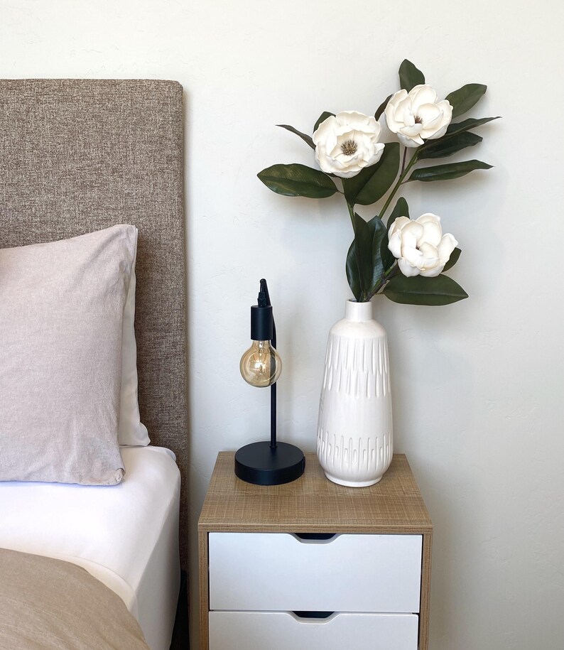 May include: A bedroom nightstand with a black lamp and a white vase holding white magnolia flowers. The nightstand has a light wood top and two white drawers. A bed with a beige headboard and pillow is in the background.