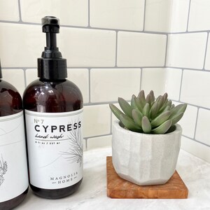 May include: Brown hand wash bottle with a black pump, labeled "No. 7 CYPRESS hand wash" and a sprig illustration. A small succulent plant in a geometric white pot sits on a wooden coaster.