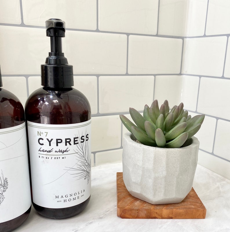 May include: Brown hand wash bottle with a black pump, labeled "No. 7 CYPRESS hand wash" and a sprig illustration. A small succulent plant in a geometric white pot sits on a wooden coaster.