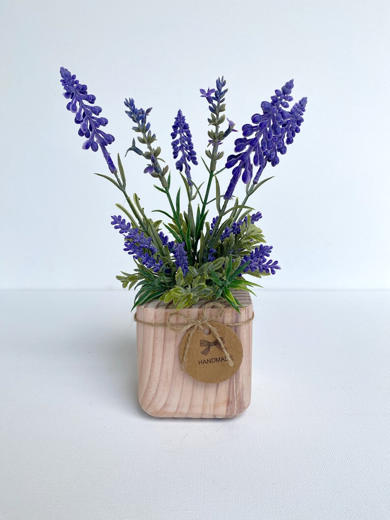 K&ouml;nnte beinhalten: Ein kleiner Holztopf mit einer lila Lavendelpflanze. Der Topf hat ein braunes Etikett mit der Aufschrift "HANDMADE".