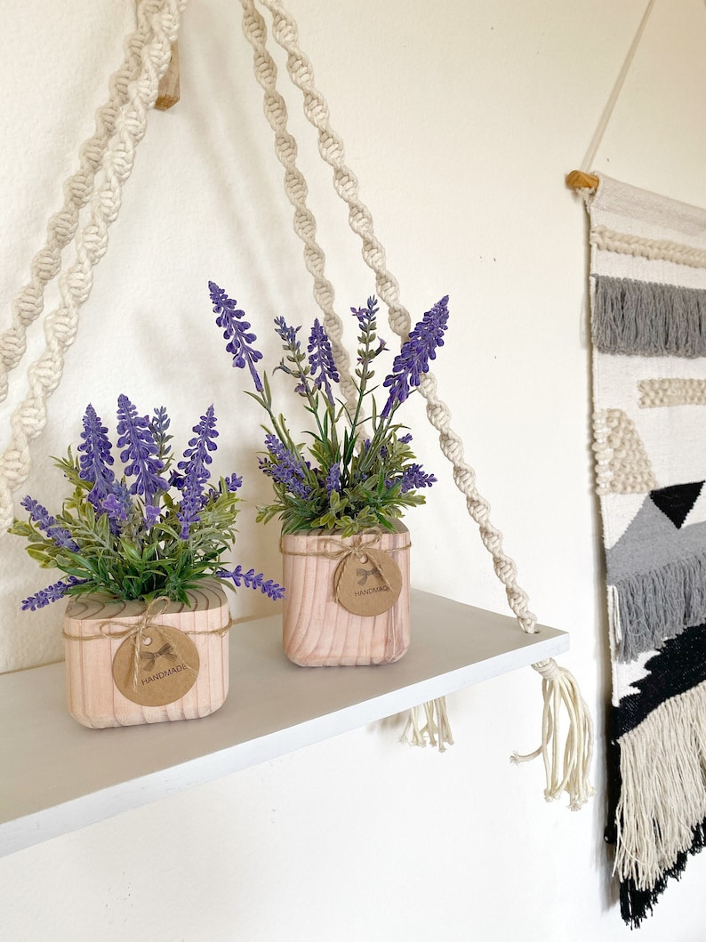 K&ouml;nnte beinhalten: Zwei Topfpflanzen mit Lavendel und lila Bl&uuml;ten in quadratischen Holzbeh&auml;ltern mit einem braunen Etikett mit der Aufschrift "Handmade". Die Beh&auml;lter stehen auf einem wei&szlig;en Regal mit einem dar&uuml;ber h&auml;ngenden Makramee-Regal.