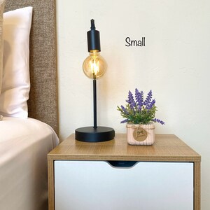 K&ouml;nnte beinhalten: Ein schwarzer Nachttisch mit zwei Schubladen, eine kleine Topfpflanze mit Lavendel und eine schwarze Tischlampe mit einer klaren Gl&uuml;hbirne. Der Text "Small" steht an der Wand.