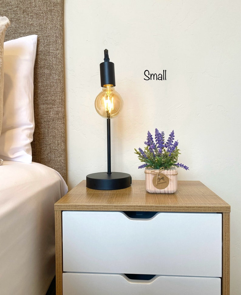 K&ouml;nnte beinhalten: Ein schwarzer Nachttisch mit zwei Schubladen, eine kleine Topfpflanze mit Lavendel und eine schwarze Tischlampe mit einer klaren Gl&uuml;hbirne. Der Text "Small" steht an der Wand.
