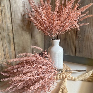 May include: Two bouquets of blush pink dried flowers, one in a white ceramic vase and the other on a white surface with a wooden bead garland and a jute tassel.