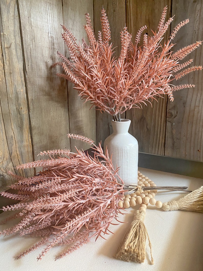 May include: Two bouquets of blush pink dried flowers, one in a white ceramic vase and the other on a white surface with a wooden bead garland and a jute tassel.