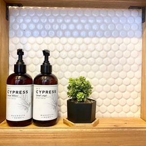 May include: Two brown bottles of hand lotion and hand wash with white labels that say "No. 7 Cypress" and "Magnolia Home". A small potted succulent plant sits on a wooden shelf with a hexagonal wooden coaster.