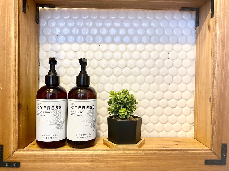 May include: Two brown bottles of hand lotion and hand wash with white labels that say "No. 7 Cypress" and "Magnolia Home". A small potted succulent plant sits on a wooden shelf with a hexagonal wooden coaster.