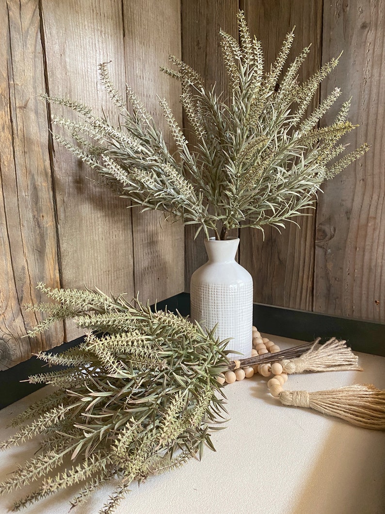 May include: Two bunches of dried, light green, feathery sprigs in white ceramic vases with a wooden bead garland and two tassels.