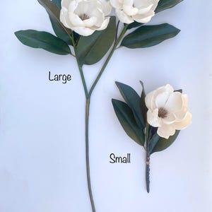 May include: Two artificial magnolia flower stems. One stem has three large white flowers and dark green leaves. The other stem has one smaller white flower and dark green leaves. The words "Large" and "Small" are written on the image.
