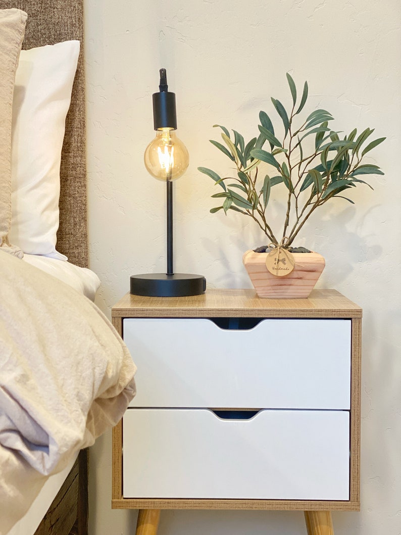 May include: A wooden nightstand with two white drawers and a black lamp with a light bulb. A potted olive tree sits on the nightstand.