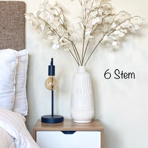 May include: White ceramic vase with a textured surface, filled with a bouquet of six white faux stems with round leaves. The vase is on a white nightstand with two drawers. The text "6 Stem" is visible in the image.