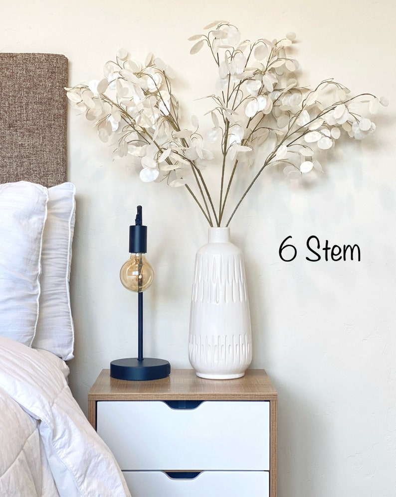 May include: White ceramic vase with a textured surface, filled with a bouquet of six white faux stems with round leaves. The vase is on a white nightstand with two drawers. The text "6 Stem" is visible in the image.