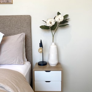 May include: A bedroom nightstand with a white and wood finish. It features two drawers and a black lamp with a lightbulb. A white vase with white flowers and green leaves sits on top. A bed with a beige headboard and bedding is visible.
