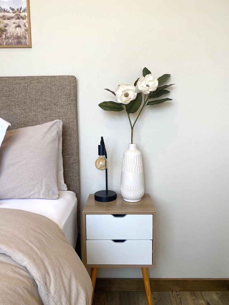 May include: A bedroom nightstand with a white and wood finish. It features two drawers and a black lamp with a lightbulb. A white vase with white flowers and green leaves sits on top. A bed with a beige headboard and bedding is visible.