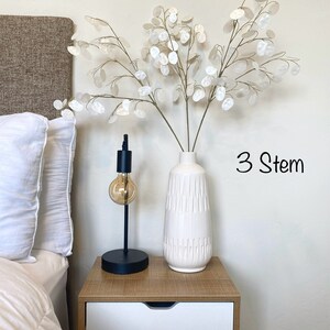 May include: Three stems of white dried flowers in a white ceramic vase with a textured design. The vase is on a wooden nightstand with a white drawer. The text "3 Stem" is written in black on the wall.