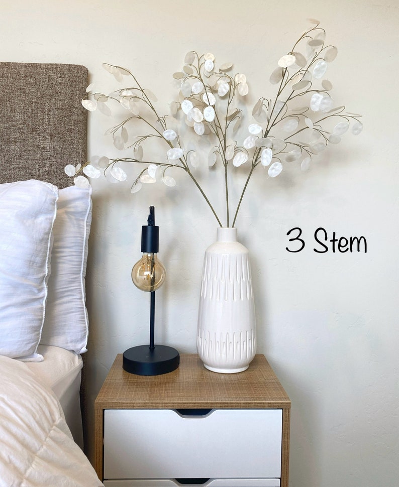 May include: Three stems of white dried flowers in a white ceramic vase with a textured design. The vase is on a wooden nightstand with a white drawer. The text "3 Stem" is written in black on the wall.