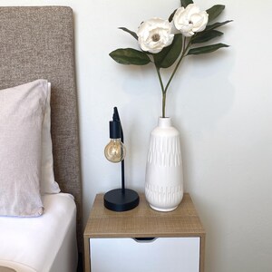 May include: A bedroom nightstand with a black desk lamp and a white vase holding white flowers and green leaves. The nightstand is made of wood and has two white drawers. A bed with a beige headboard and white bedding is in the background.