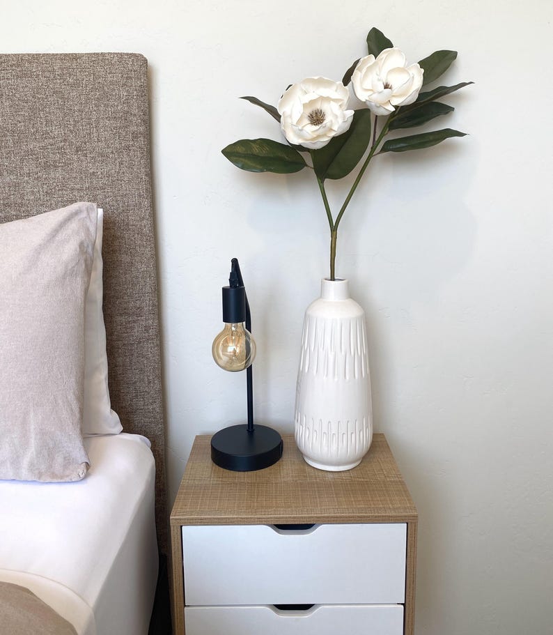 May include: A bedroom nightstand with a black desk lamp and a white vase holding white flowers and green leaves. The nightstand is made of wood and has two white drawers. A bed with a beige headboard and white bedding is in the background.