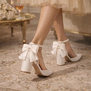 May include: Cream-coloured block heel pumps featuring large satin bows and pearl ankle straps. The shoes have a pointed toe and a chunky heel. The background includes a patterned rug and a table with a glass of wine.