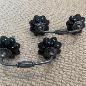 May include: Four black, flower-shaped cabinet pulls with silver-toned hardware. The pulls have a glossy finish and are arranged on a neutral, textured surface. The hardware includes a decorative centre and a curved handle.