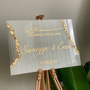May include: A rectangular sign with gold text and accents, displayed on a wooden easel. The sign features the names "S&uuml;meyye & Enes" and the date "18.05.2023". Gold leaf accents the edges. The sign has text in a foreign language.