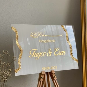 May include: A rectangular wedding welcome sign with gold text and accents. The sign reads "Hoşgeldiniz Tugce & Eren 09.08.2023" in gold lettering. The sign is displayed on a wooden easel.