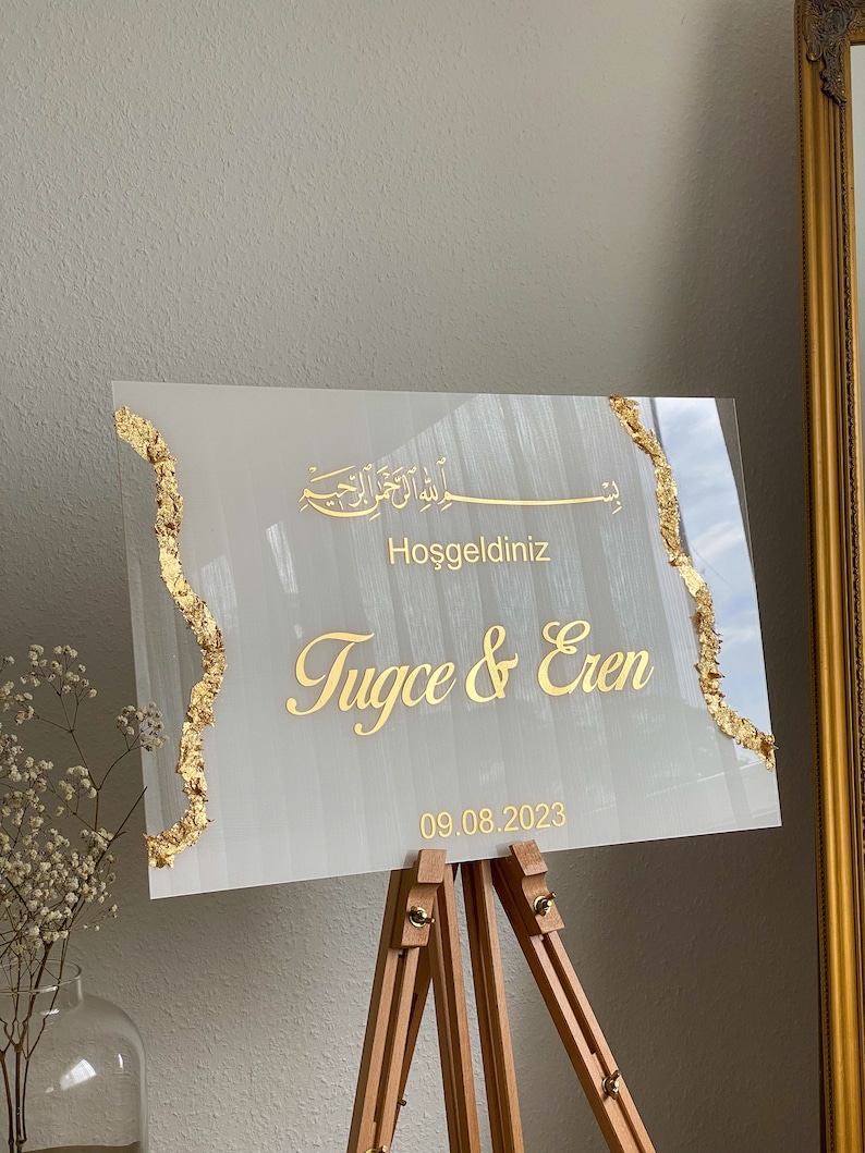 May include: A rectangular wedding welcome sign with gold text and accents. The sign reads "Hoşgeldiniz Tugce & Eren 09.08.2023" in gold lettering. The sign is displayed on a wooden easel.